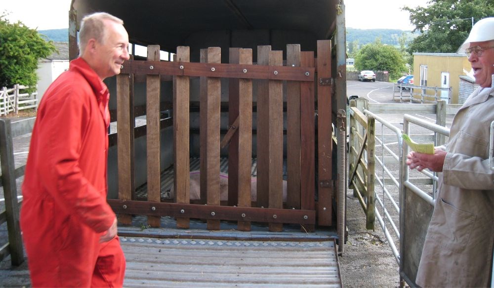 Two men wearing overalls standing in front of a small livestock trailer.